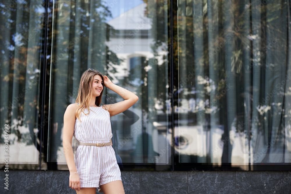 Fototapeta premium Young pretty blond woman, wearing white stripy short overall and sunglasses, standing near big glass building. Three-quarter portrait of slim girl posing for picture in resort. Vacation leisure time