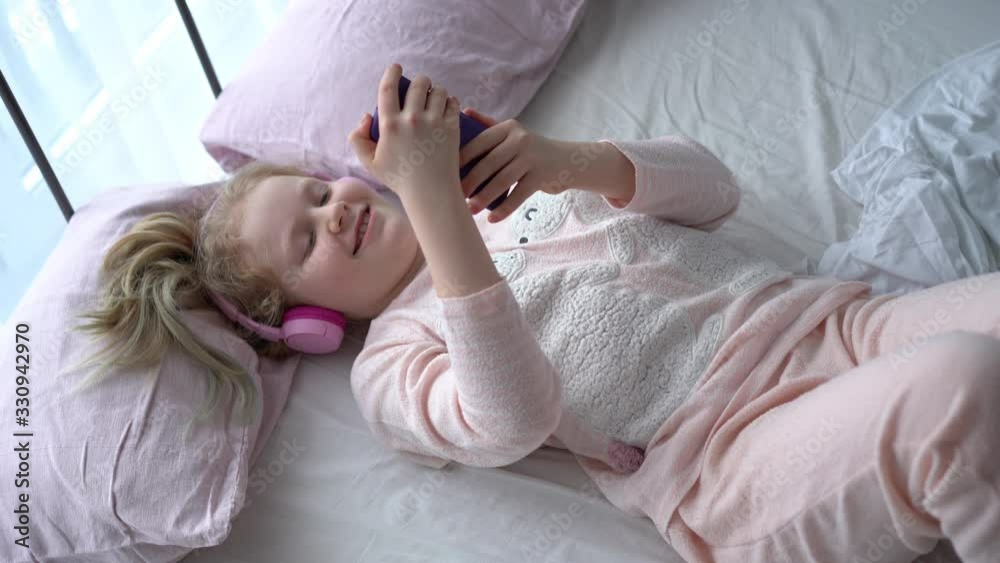 modern life of generation Z. teenage girl in pajamas and headphones in the room on the bed listens to music from a smartphone. modern life of generation Z. teenage girl in pajamas and headphones in the room on the bed listens to music from a smartphone.