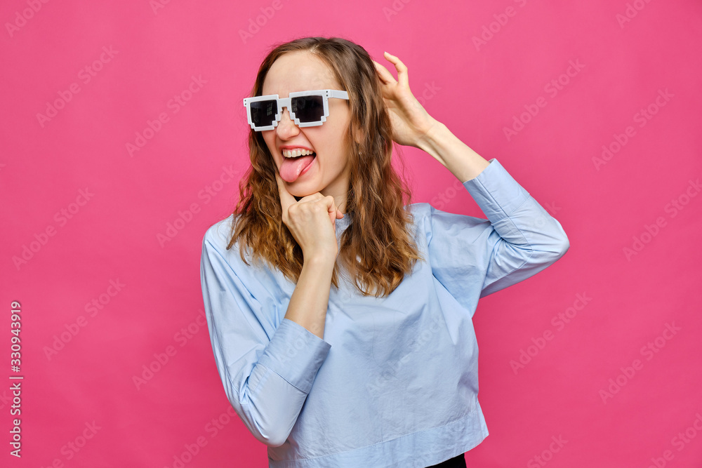 Stylish caucasian girl in a pale blue t-shirt and 8-bit glasses shows tongue and grimaces on a pink background.