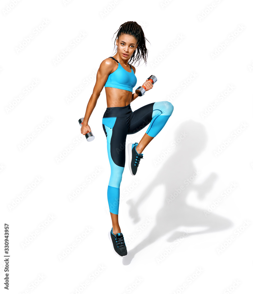 Sporty girl jumping with dumbbells. Photo of african american girl in fashionable sportswear on white background. Dynamic movement. Full length. Sports and healthy lifestyle