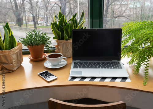 Laptop with empty screen with plants, cup of coffee and smartphone near the window on a wooden office desk. Business and workplace in cafe. Freelancer and work in coffee shop.