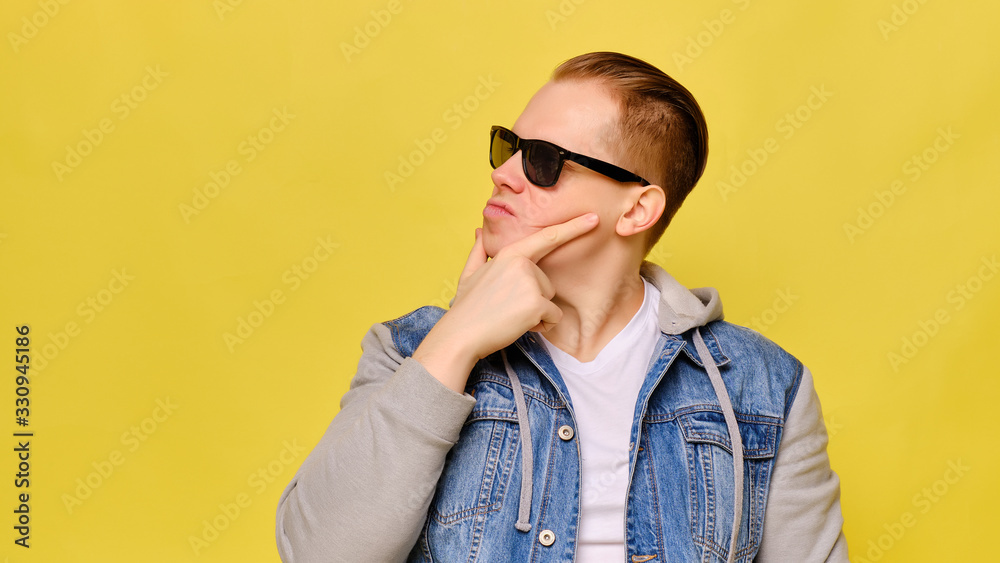 Stylish Caucasian man in a jeans and sunglasses on a yellow background. Copy space. Abstractly looks at the object with doubt. Stylish Caucasian man in a jeans and sunglasses on a yellow background. Copy space. Abstractly looks at the object with doubt.