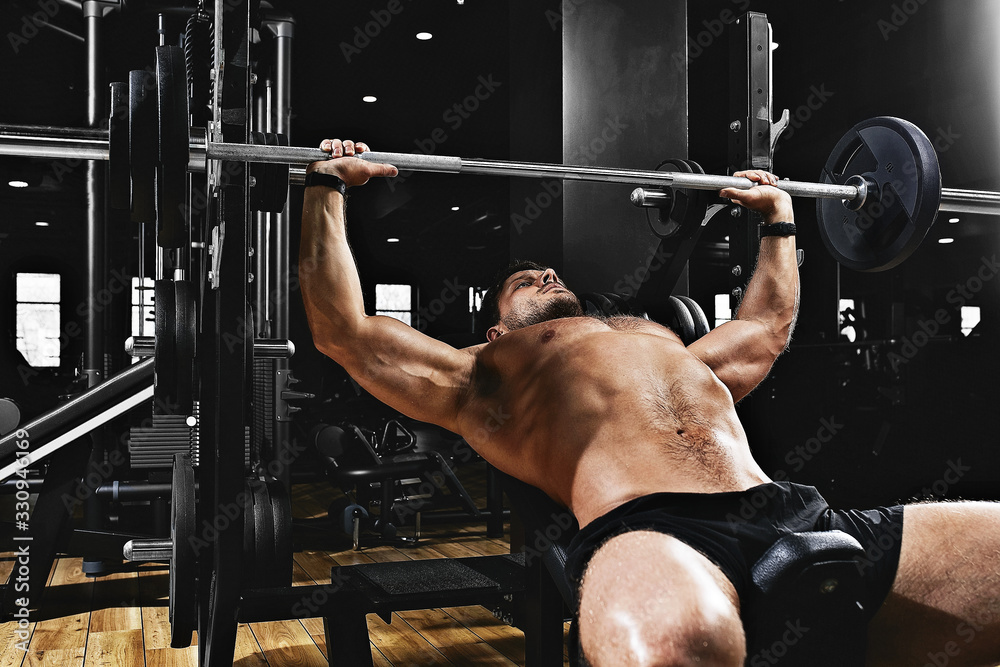 Fototapeta premium Young muscular man lifting a barbell bench press in the gym. Sport, movement, life. The concept of a healthy lifestyle, regular training, development of the pectoral muscles