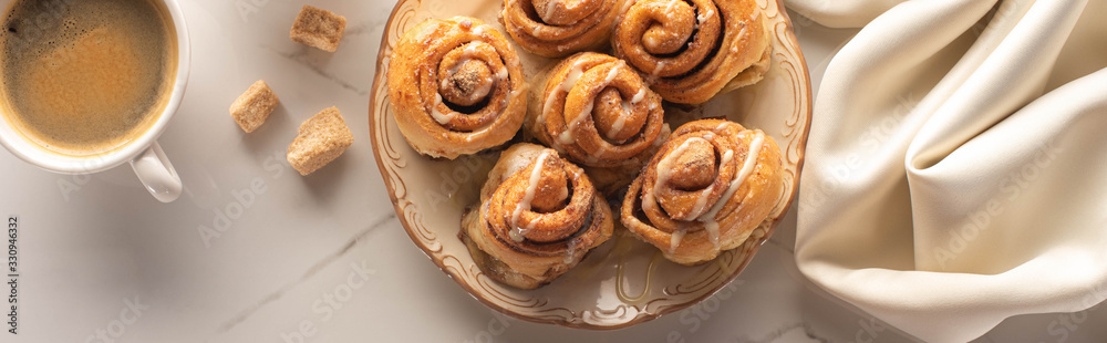 top view of fresh homemade cinnamon rolls on marble surface with cup of coffee, condensed milk and silk cloth, panoramic shot top view of fresh homemade cinnamon rolls on marble surface with cup of coffee, condensed milk and silk cloth, panoramic shot