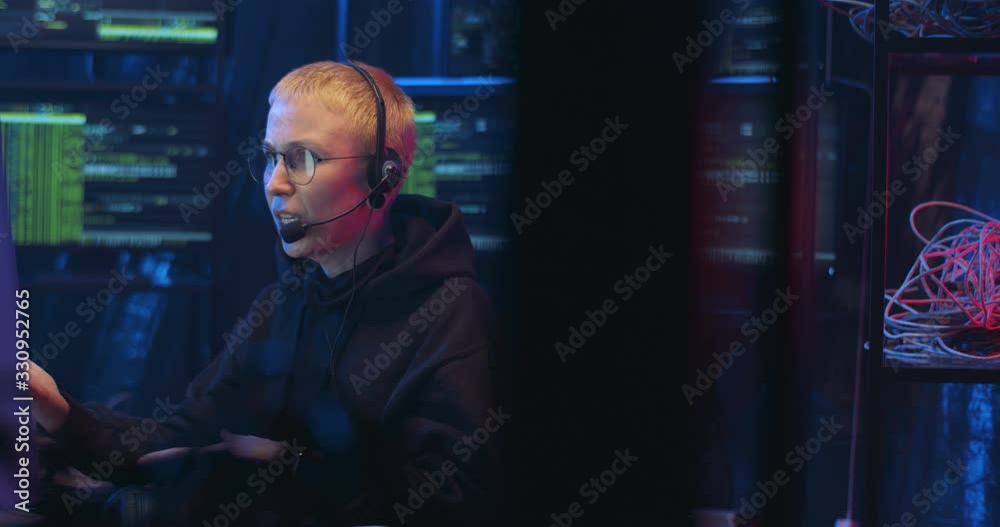 Caucasian woman IT orker in headset speaking in mic while working at compuer screen in call monitoring center in dark room at night. Female cyber security policewoman talking with client.