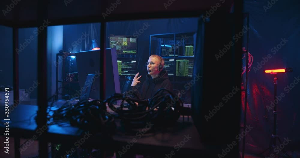 Caucasian woman in headset with mic talking while having call with client in dark room with monitors. Female worker of IT call-center in front of computer screen speaking and browsing online. Caucasian woman in headset with mic talking while having call with client in dark room with monitors. Female worker of IT call-center in front of computer screen speaking and browsing online.