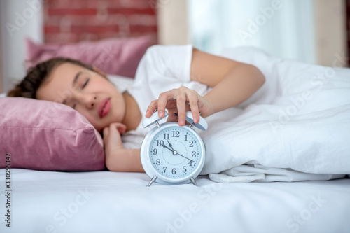 Sleeping teenager girl in bed, hand on the alarm clock.