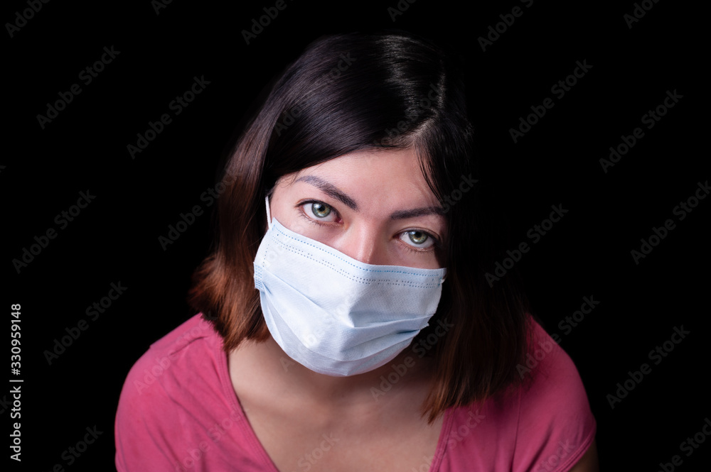 Brunette young girl with green eyes with a white surgical face mask from a coronavirus pandemic disease infection COVID-19 isolated on black background Brunette young girl with green eyes with a white surgical face mask from a coronavirus pandemic disease infection COVID-19 isolated on black background