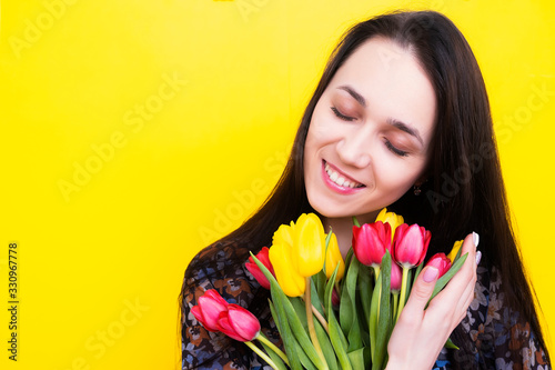 Portrait of a brunette woman with a bouquet of tulips on a yellow background. Birthday girl with flowers. Happy woman with a bouquet of flowers smiling closing her eyes.