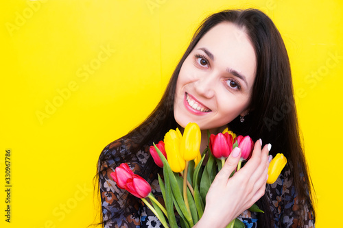 A birthday girl with a bouquet of tulips looks out of the frame and smiles. Happy brunette with a bouquet of flowers. Florist with flowers on a yellow background.