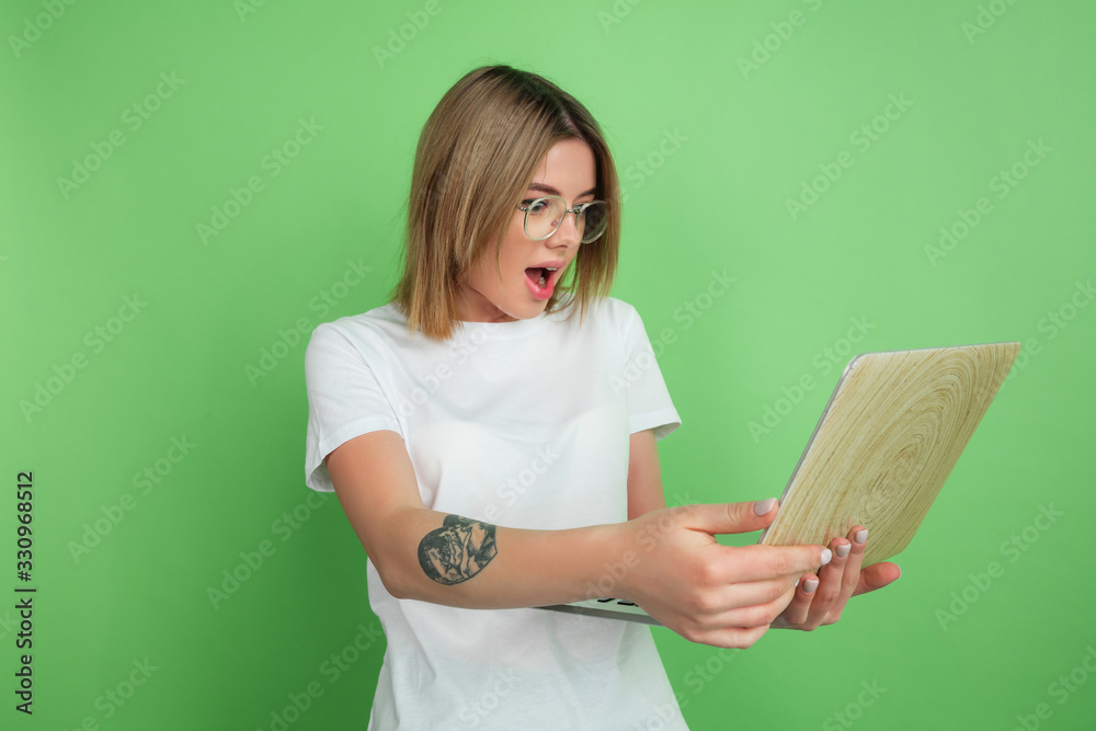 Using laptop emotional. Caucasian young woman's portrait isolated on green studio background. Beautiful female model in white shirt. Concept of human emotions, facial expression, sales, ad, youth.