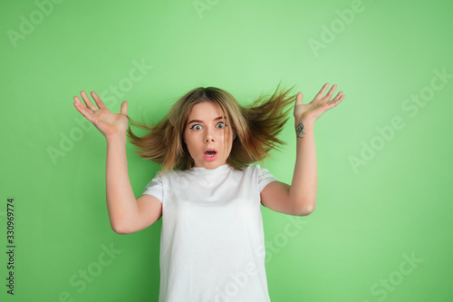 Wondered. Caucasian young woman's portrait isolated on green studio background. Beautiful female model in white shirt. Concept of human emotions, facial expression, sales, ad, youth. © master1305