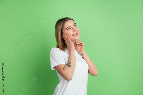 Dreaming, idea. Caucasian young woman's portrait isolated on green studio background. Beautiful female model in white shirt. Concept of human emotions, facial expression, sales, ad, youth.