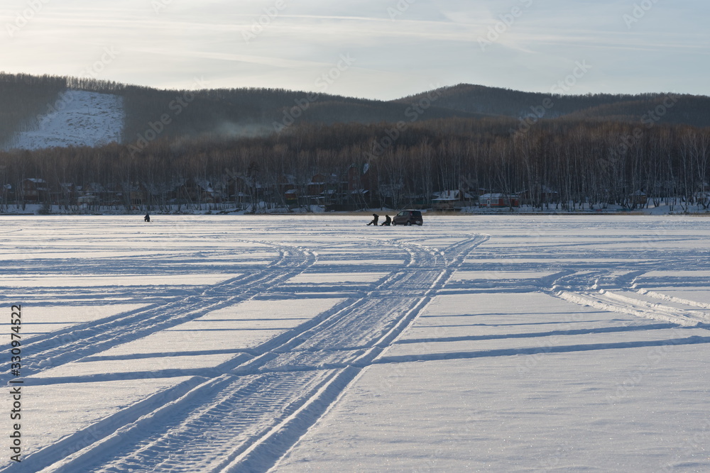 Traces from the wheels of cars lead along the snowy plain of the lake to the fishermen against the background of the coast. Traces from the wheels of cars lead along the snowy plain of the lake to the fishermen against the background of the coast.