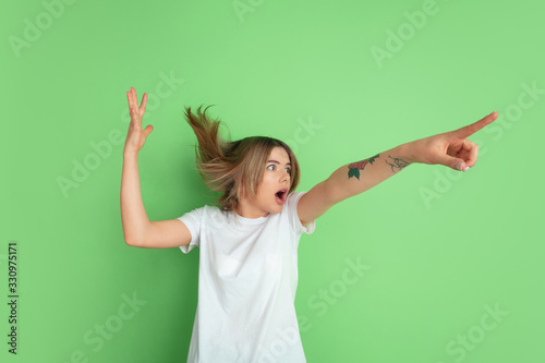 Shocked pointing up. Caucasian young woman's portrait isolated on green studio background. Beautiful female model in white shirt. Concept of human emotions, facial expression, sales, ad, youth. © master1305