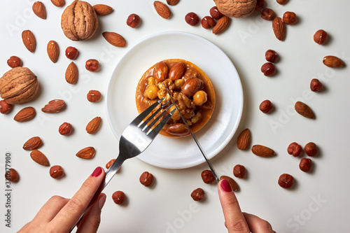 Woman hands with knife and fork cutting mini tart cupcake with walnuts, almonds and hazelnuts on plate with knife and fork. Close up. Top view