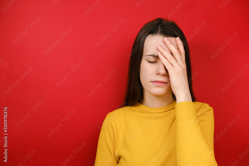 Frustrated woman holding hands on forehead being depressed regretting what she done having headache, wears yellow sweater, stands over red background. Negative human emotions and feelings concept Frustrated woman holding hands on forehead being depressed regretting what she done having headache, wears yellow sweater, stands over red background. Negative human emotions and feelings concept