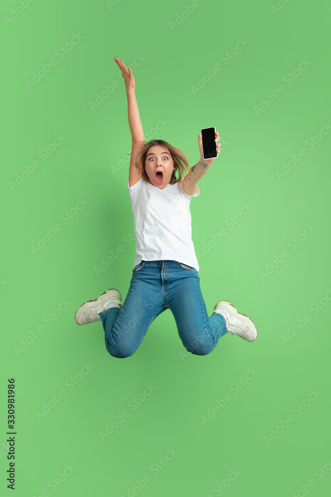 Showing phone screen in jump. Caucasian young woman's portrait isolated on green studio background. Beautiful female model in white shirt. Concept of human emotions, facial expression, sales, ad Showing phone screen in jump. Caucasian young woman's portrait isolated on green studio background. Beautiful female model in white shirt. Concept of human emotions, facial expression, sales, ad