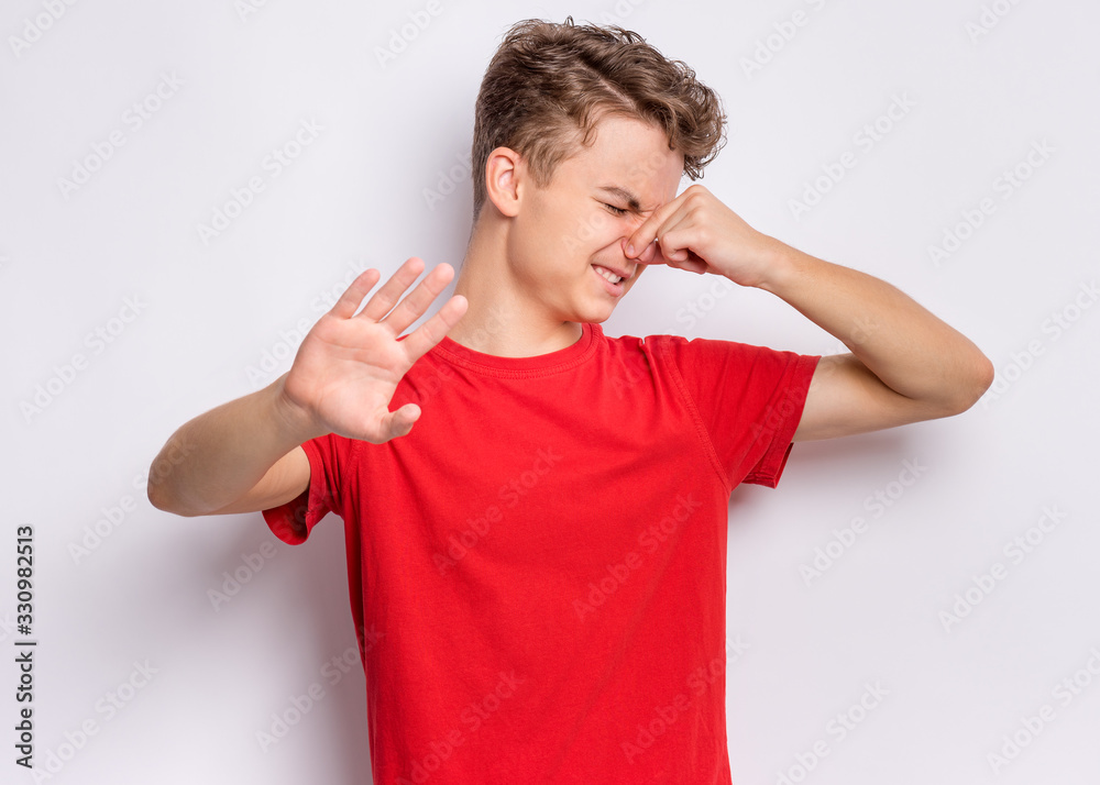 Portrait of teen boy covering nose with hand and doing stop gesture. Child covers nose with gesture smells bad. Teenager shows that something stinks and shows stop sign with palm on grey background. Portrait of teen boy covering nose with hand and doing stop gesture. Child covers nose with gesture smells bad. Teenager shows that something stinks and shows stop sign with palm on grey background.