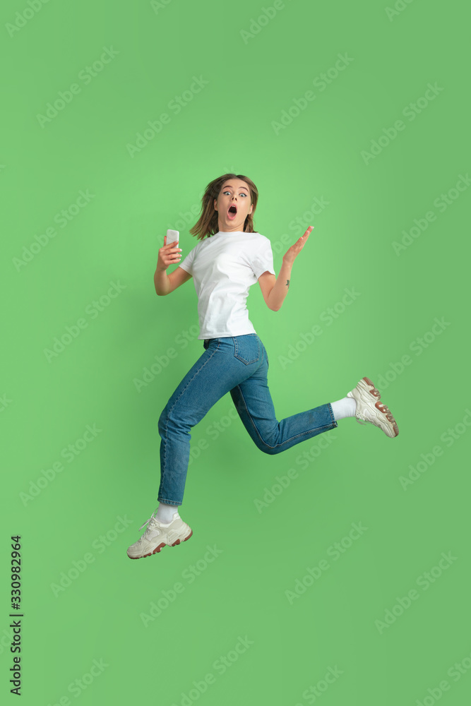 Shocked using phone in jump. Caucasian young woman's portrait isolated on green studio background. Beautiful female model in white shirt. Concept of human emotions, facial expression, sales, ad, youth Shocked using phone in jump. Caucasian young woman's portrait isolated on green studio background. Beautiful female model in white shirt. Concept of human emotions, facial expression, sales, ad, youth