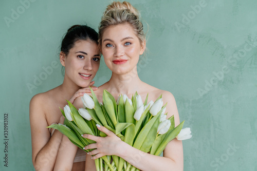 Beauty portrait of young gorgeous attractive blonde and brunette serious women posing naked isolated over beige wall background with green nature leaf plant.