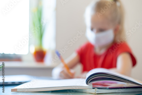 Blurred schoolgirl in medical mask studying at home, doing school homework, writing in notebook. Reading training books on table.coronavirus quarantine. Distance learning online education
