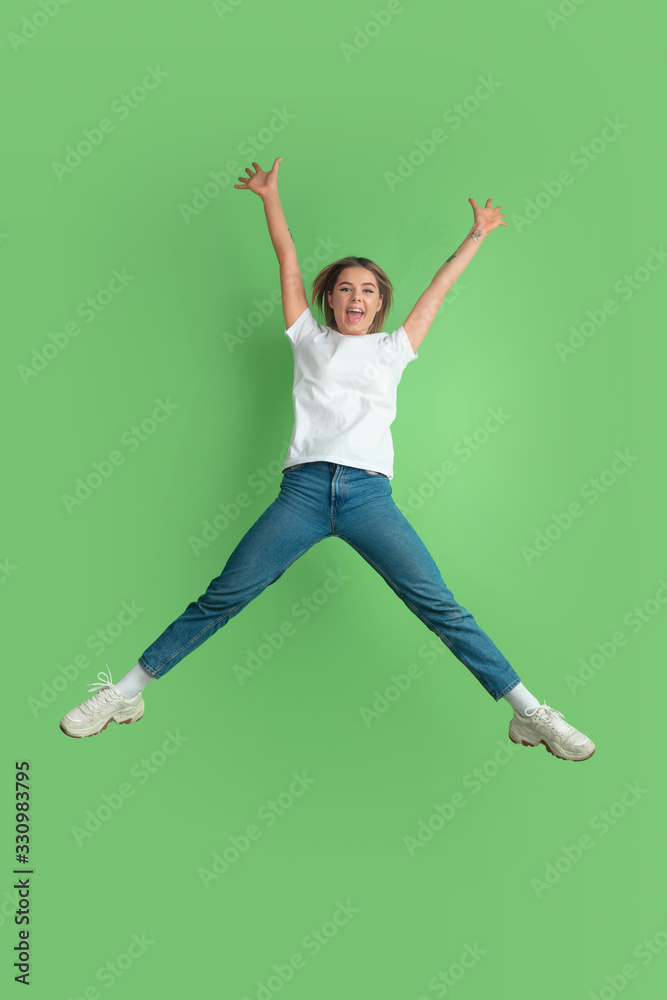 Crazy happy in jump. Caucasian young woman's portrait isolated on green studio background. Beautiful female model in white shirt. Concept of human emotions, facial expression, sales, ad, youth.
