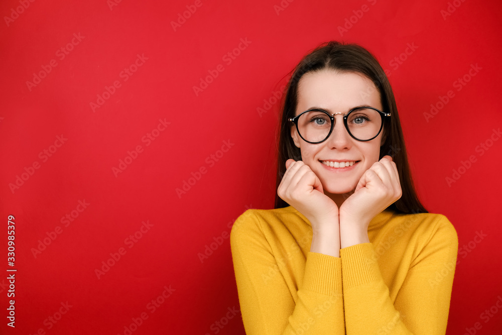 Pleasant looking friendly cute young woman in glasses holds chin with both hands and smiles positively, shows white teeth, wears sweater, stands over red background. People , good emotions concept Pleasant looking friendly cute young woman in glasses holds chin with both hands and smiles positively, shows white teeth, wears sweater, stands over red background. People , good emotions concept