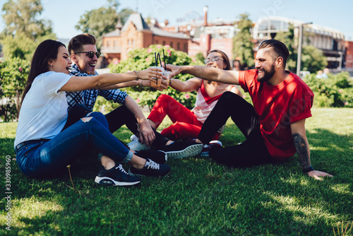 Overjoyed students in casual clothing celebrating holidays and toasting while resting on green grass