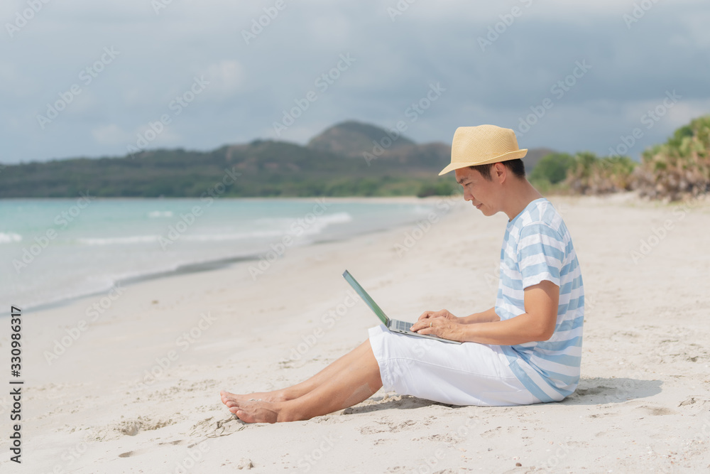 Asian man on holiday wearing a hat sit on the beach and working with a laptop.