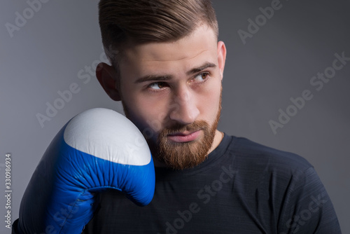 Portrait of handsome bearded brutal guy in sportswear standing in a combat pose, holding raised hands near his face, in boxing gloves. On a gray background. Sport lifestyle. © Алексей Доненко
