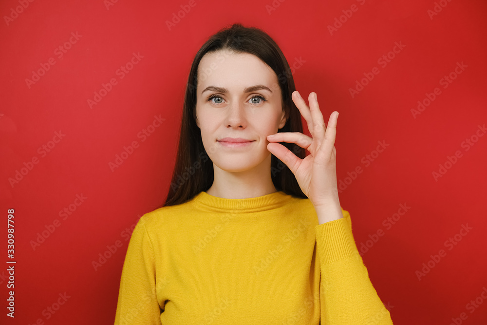 Sly smiling young woman looks mysteriously, aside zips her mouth shut, promises to keep secret, holds fingers near cheek, hides information, dressed in yellow sweater, isolated on red background Sly smiling young woman looks mysteriously, aside zips her mouth shut, promises to keep secret, holds fingers near cheek, hides information, dressed in yellow sweater, isolated on red background