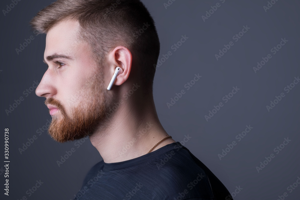 Fototapeta premium Zbliżenie portret młodego brodatego faceta dwudziestu pięciu lat, stojąc w profilu. W bezprzewodowych białych słuchawkach. Sportowy styl. Zdjęcie promocyjne słuchawek sportowców