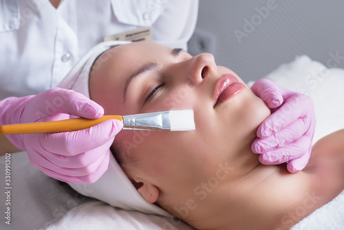 close-up, hands of a professional cosmetologist doctor, using a tool, performs the application of cream and oil on the skin of the face of a young patient. In the beauty salon