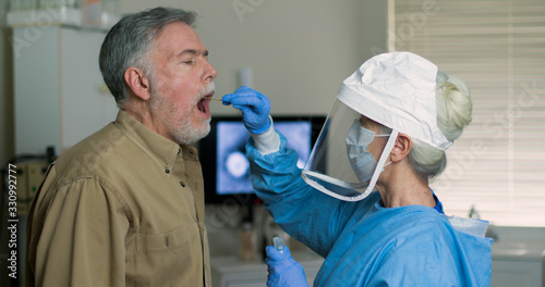 A mature Caucasian man in a clinical setting being swabbed by a healthcare worker in protective garb to determine if he has contracted the coronavirus or COVID-19.. photo