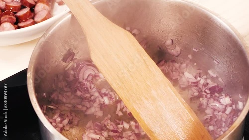 Oil is slowly poured into a large pan with finely chopped red onions and a wooden spatula.