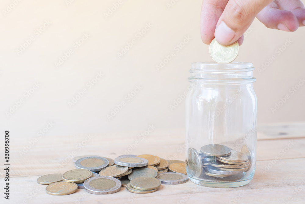 Fototapeta premium The hand hold coin and putting saving coins into glass with a pile of coinson wooden table - Investment, Business, Finance and Banking Concept