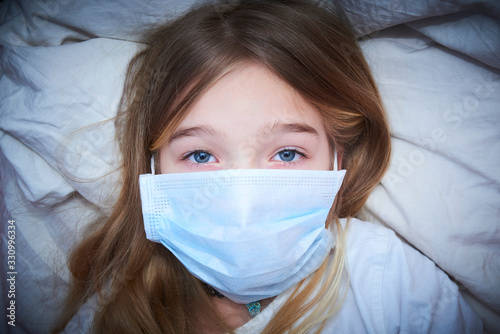 Sick girl lying in bed with protection mask on face against infection. Selective focus