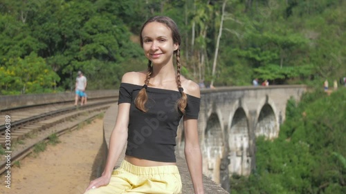 cheerful girl with braids sits on historical bridge with rails against magnificent wild forest slow motion closeup. Concept environment travel lifestyle photo