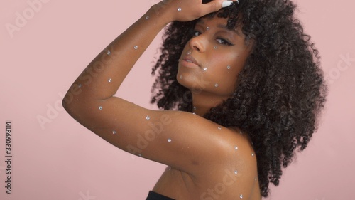 Mixed race black woman with curly hair covered by crystal makeup dancing on a pink background