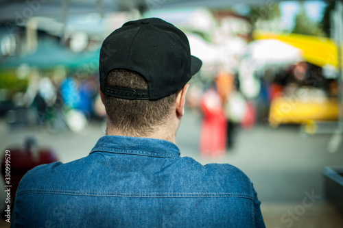 Canvas Print A large build man is seen from behind in selective focus, wearing blue denim shirt and black baseball cap at a street market with blurry stalls in background