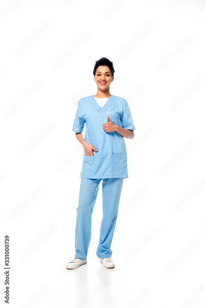 Full length view of african american nurse with hand in pocket and thumb up smiling and looking at camera on white Full length view of african american nurse with hand in pocket and thumb up smiling and looking at camera on white