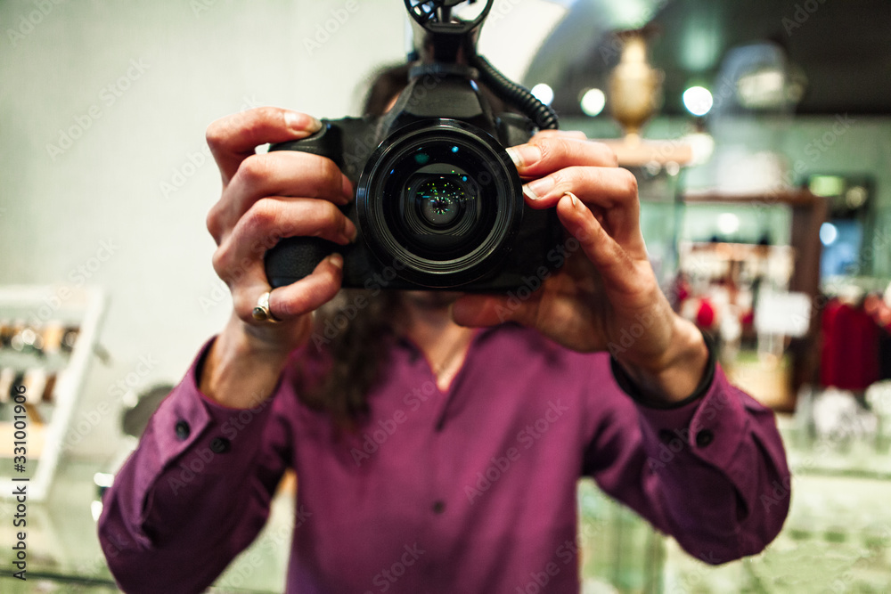 A close up and front view in selective focus of a man filming B roll, holding camera freely without tripod, with DSLR camera and lens reflections.