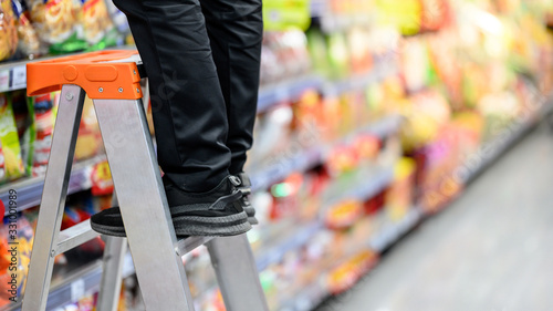 Male storekeeper legs standing on aluminum stair putting products on shelves in grocery store or supermarket. Worker filling foods stock for groceries business. photo