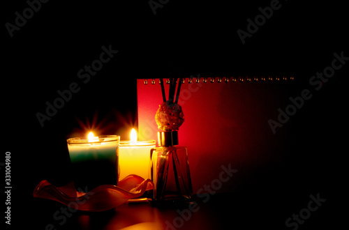 The candles in two glass bottles illuminate beside the diffuser, set in front of the red book cover on a black background.