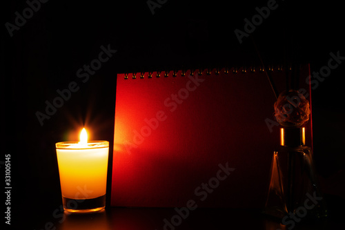 The candle light in the glass shines on the side, the red book cover on a black background, with a reed diffuser on the side.