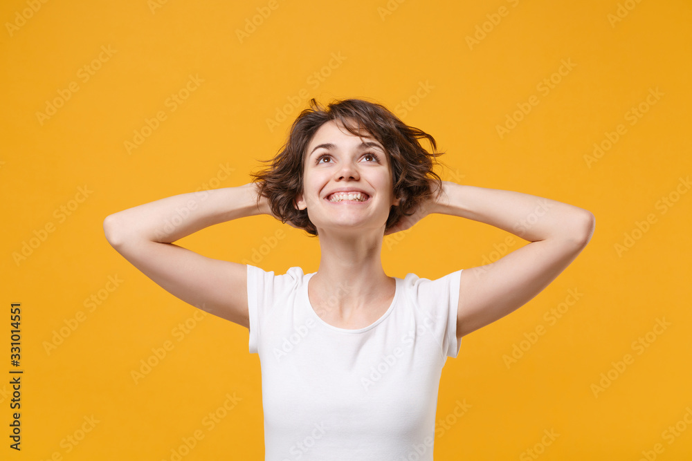 Smiling young brunette woman girl in white t-shirt posing isolated on yellow orange wall background studio portrait. People lifestyle concept. Mock up copy space. Looking up with hands behind head. Smiling young brunette woman girl in white t-shirt posing isolated on yellow orange wall background studio portrait. People lifestyle concept. Mock up copy space. Looking up with hands behind head.