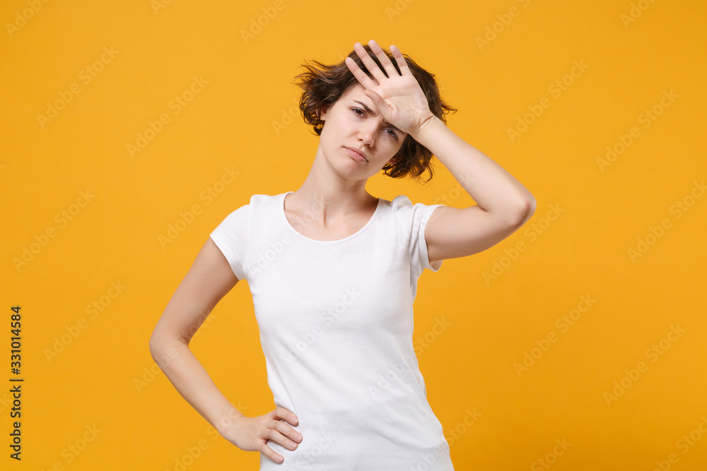 Tired young brunette woman girl in white t-shirt posing isolated on yellow orange wall background studio portrait. People sincere emotions lifestyle concept. Mock up copy space. Put hand on head.