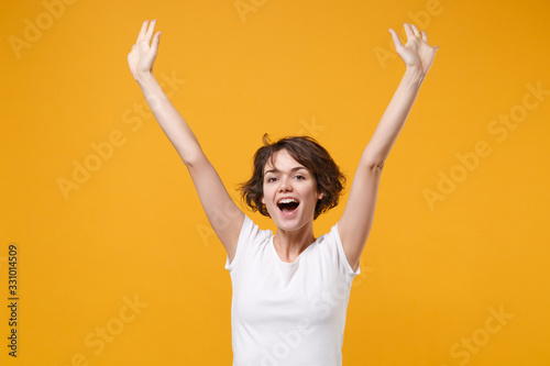 Cheerful young brunette woman girl in white t-shirt posing isolated on yellow orange background studio portrait. People sincere emotions lifestyle concept. Mock up copy space. Rising spreading hands.