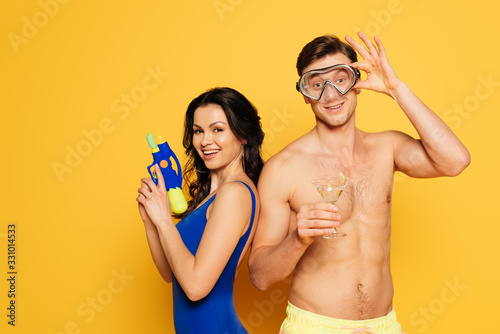 cheerful man in diving mask holding glass of cocktail near happy woman with water gun on yellow background